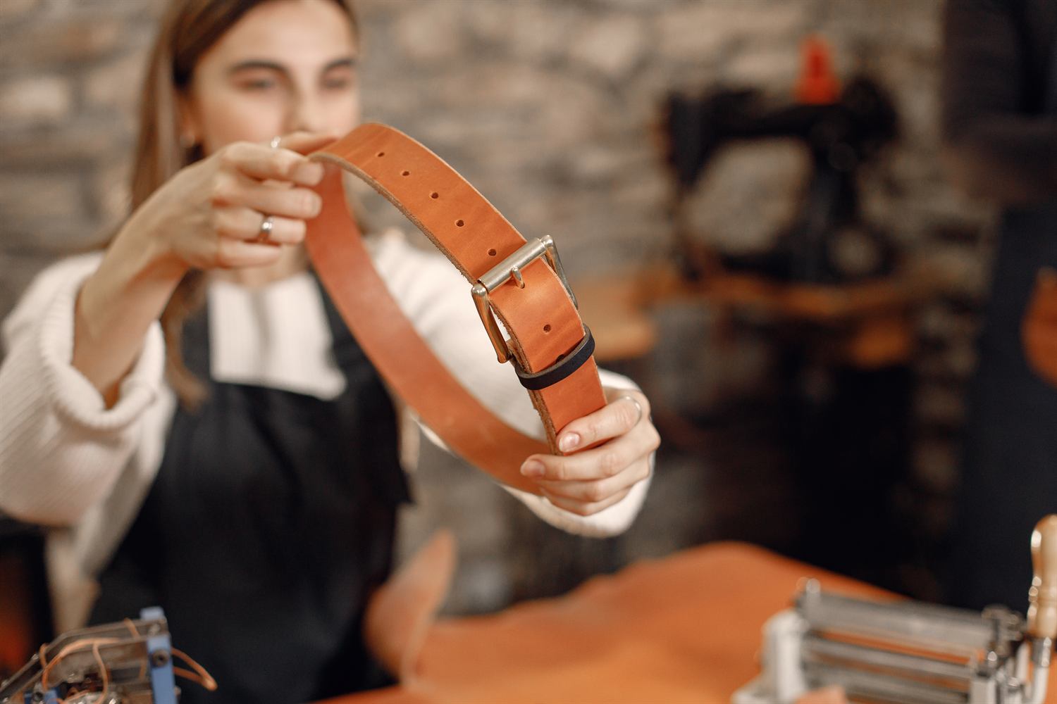 A woman in an apron holding up a tan leather belt in a workshop