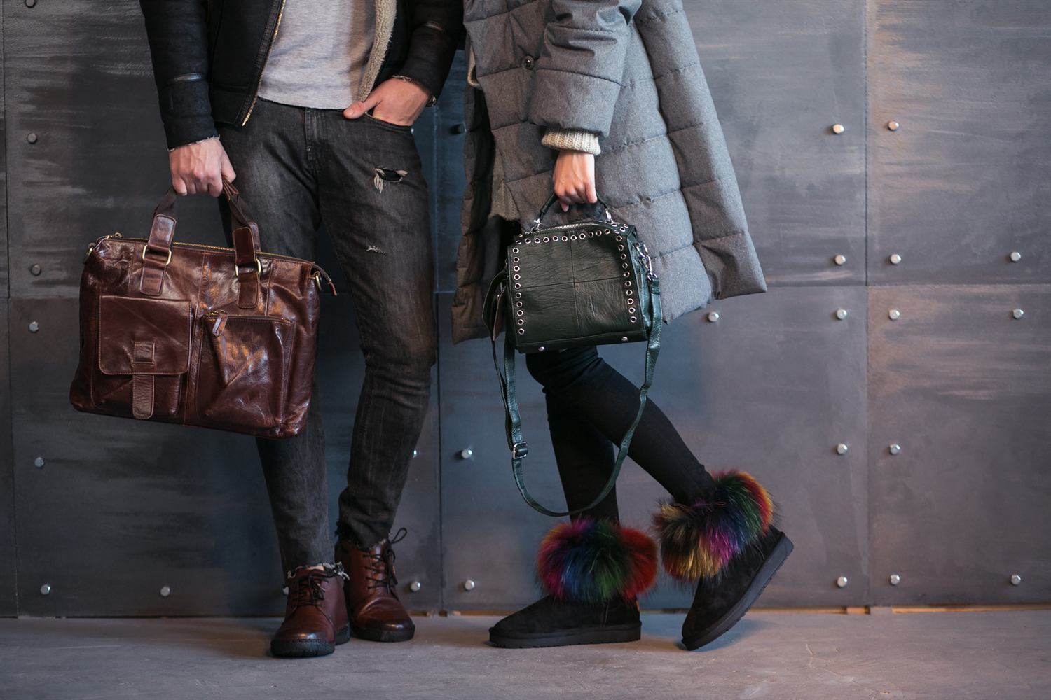 Two people standing against a metal wall, one holding a brown leather work bag and one a dark green leather shoulder bag